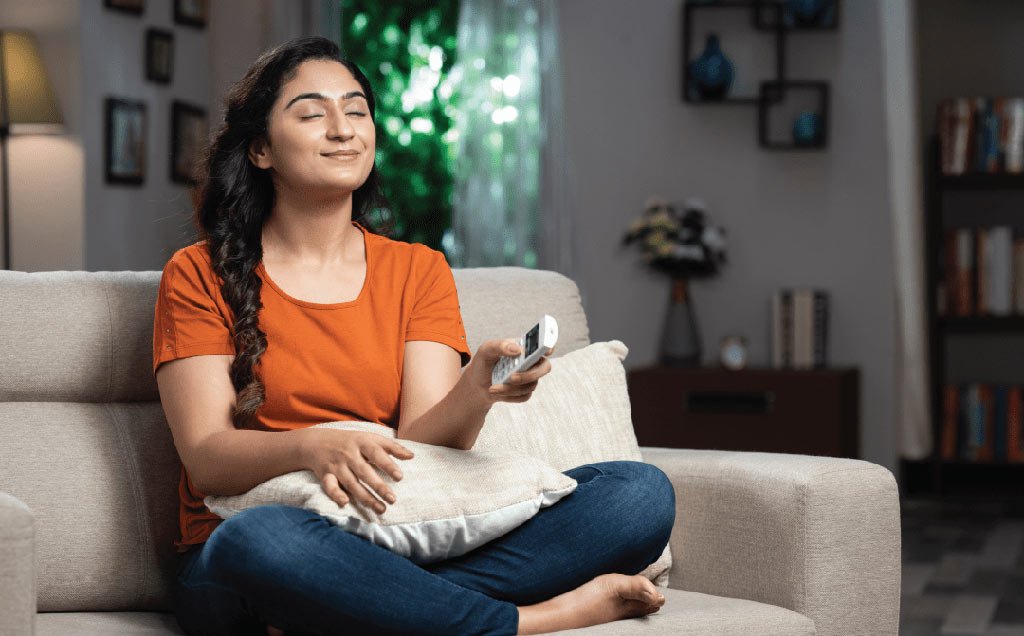 Woman sitting comfortably on a couch with a remote control, enjoying clean and refreshing air in her home, highlighting the importance of indoor air quality for a healthy living environment.